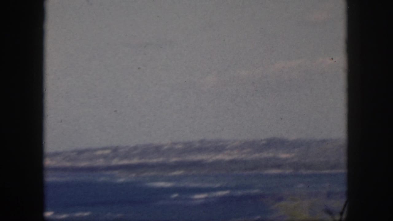 1959 Oahu shoreline footage with distant hills, blue water, scattered surf lines, and dark window-frame borders.