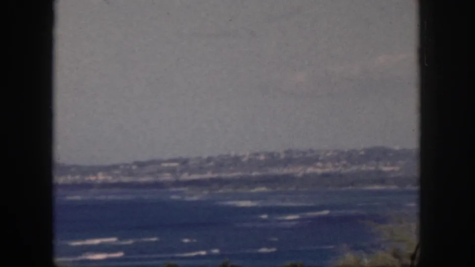 1959 Oahu ocean view with a dark window frame, hazy sky, distant shoreline, and white surf across blue water.