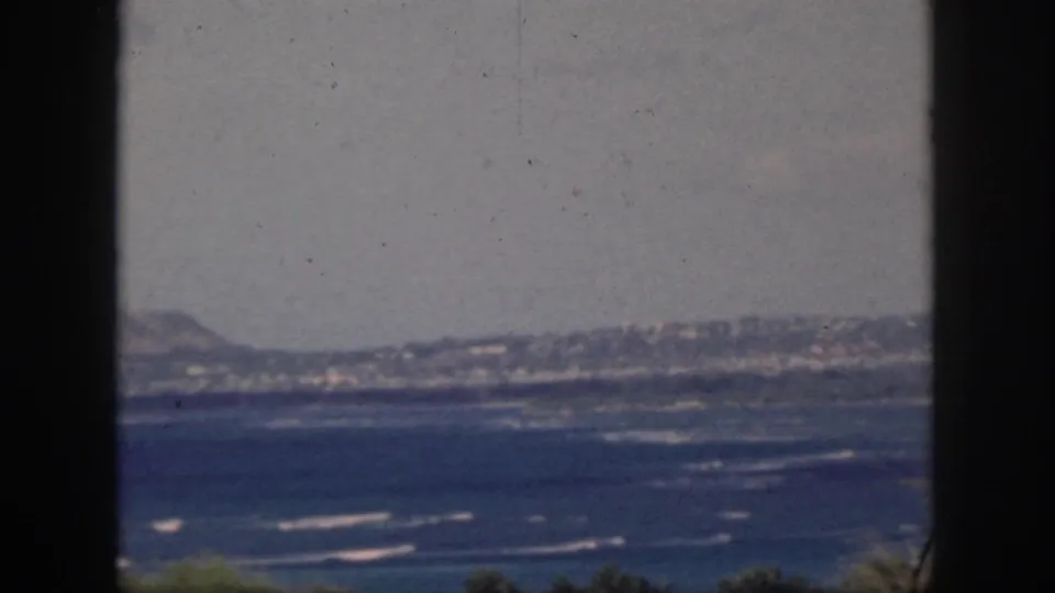 1959 Oahu coastal landscape showing blue ocean, white surf, distant hills, dark frame edges, and faint foreground foliage.