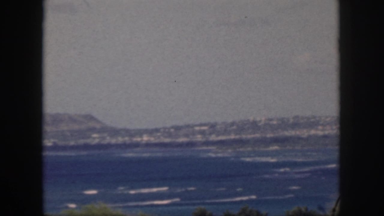 1959 Oahu coastal view with blue water, white surf lines, distant hills, and a dark foreground frame.