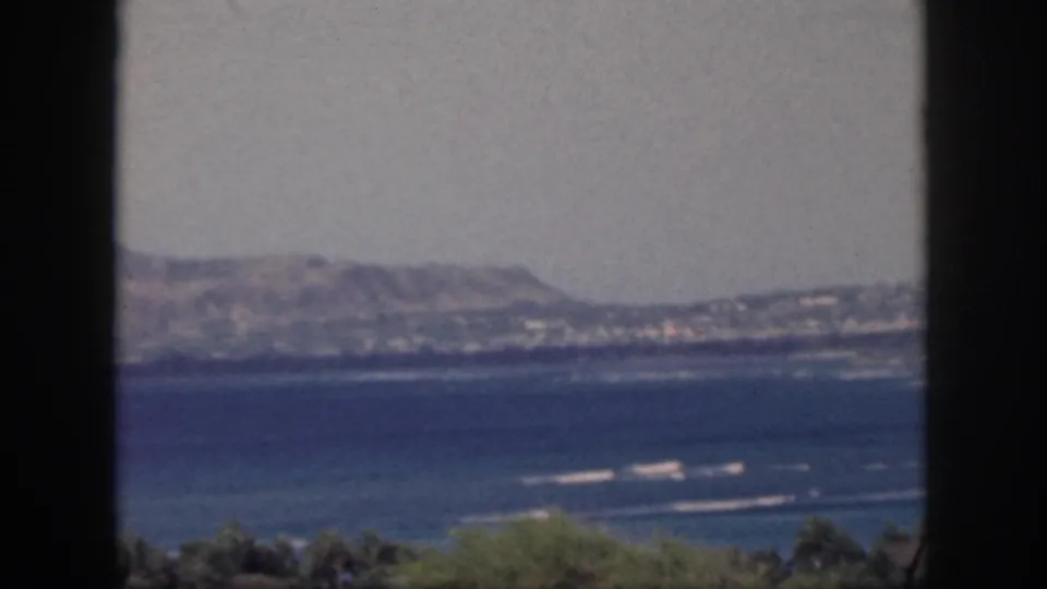 Vintage 8mm view of Oahu coast with distant hills, blue ocean, white surf streaks, and a dark frame at left.