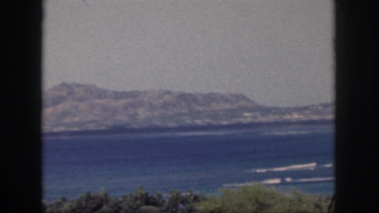 1959 Oahu home-movie view of blue ocean, surf lines, and distant hills framed by dark vertical window mullions.