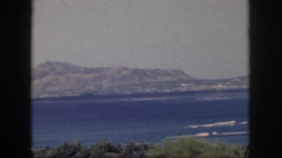1959 Oahu home-movie view of blue ocean, surf lines, and distant hills framed by dark vertical window mullions.
