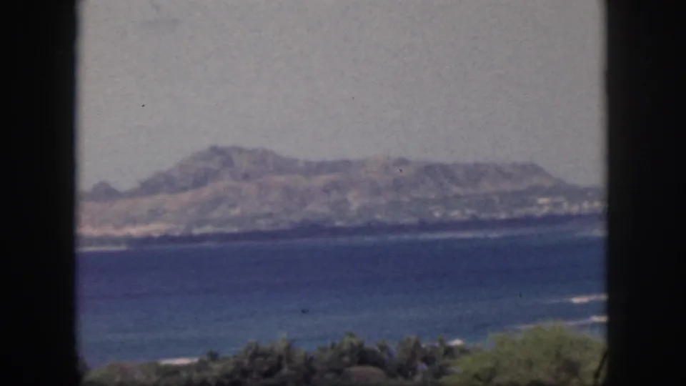 1959 Oahu coastal view with a hazy hilly shoreline, blue water, low vegetation, and dark window framing in the foreground.