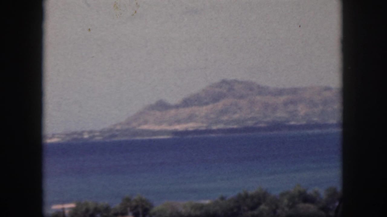 1959 Oahu home-movie frame showing a distant island or coastline across blue water through a dark window frame.