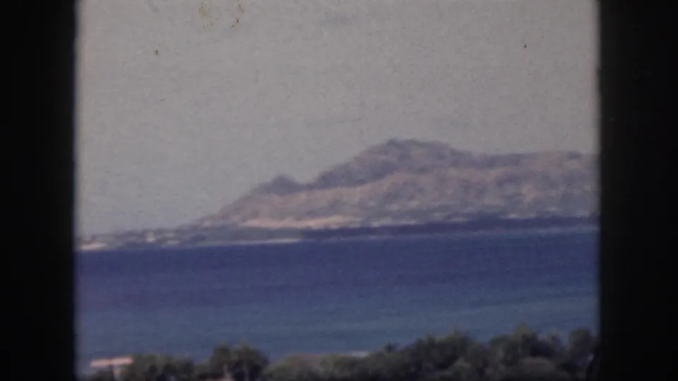 1959 Oahu home-movie frame showing a distant island or coastline across blue water through a dark window frame.