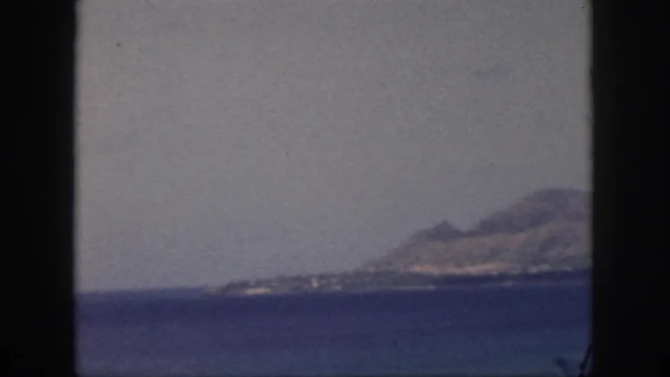 1959 Oahu home-movie frame showing a distant mountain coastline across blue water through black window edges.