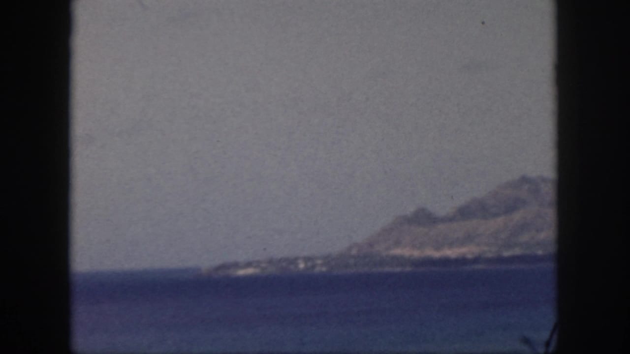 1959 Oahu home-movie view of a hazy mountain ridge across a calm bay, seen through dark vertical frame edges.