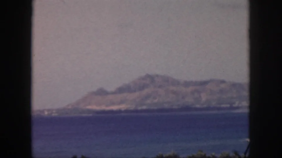 1959 Oahu 8mm frame showing a soft mountain range beyond blue bay water, with dark frame edges and faint foreground vegetation.