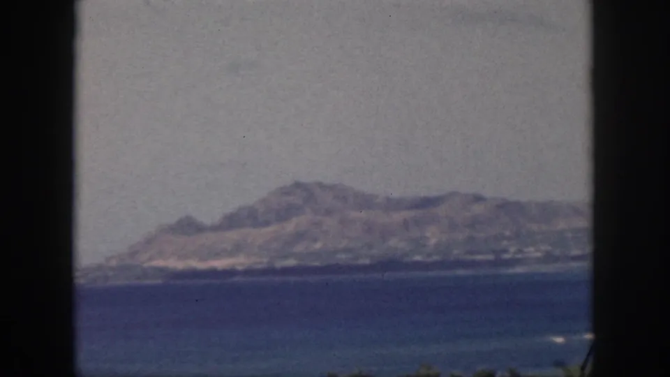Late-1950s Oahu home-movie frame of a mountain ridge across a calm bay, bordered by dark window-like edges.