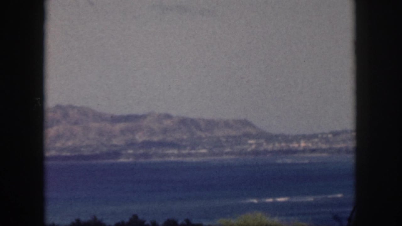 1959 Oahu home-movie view of distant coastal mountains across blue water, framed by dark vertical window edges.