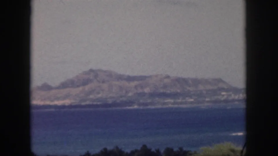 Soft 8mm view of Oahu coastal mountains beyond blue ocean, with dark framing and faint foreground vegetation.