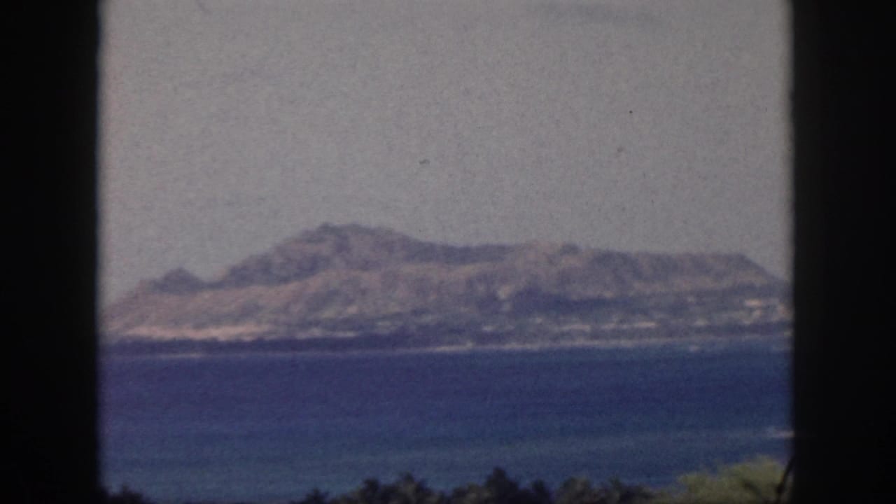1959 Oahu 8mm frame showing a hazy mountain ridge across a calm bay, with dark window or doorway edges in the foreground.