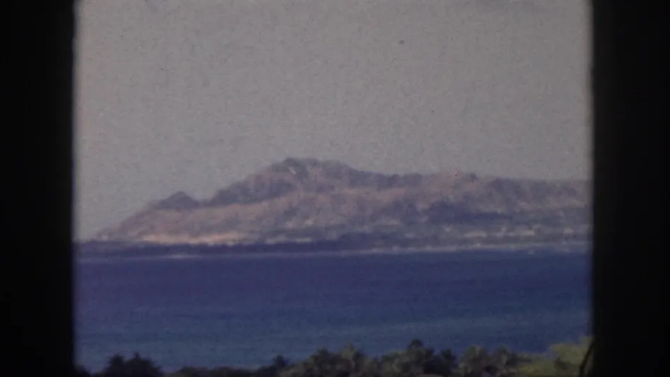 Oahu home-movie frame from 1959 showing a distant mountain and shoreline across deep blue water, bordered by dark foreground edges.