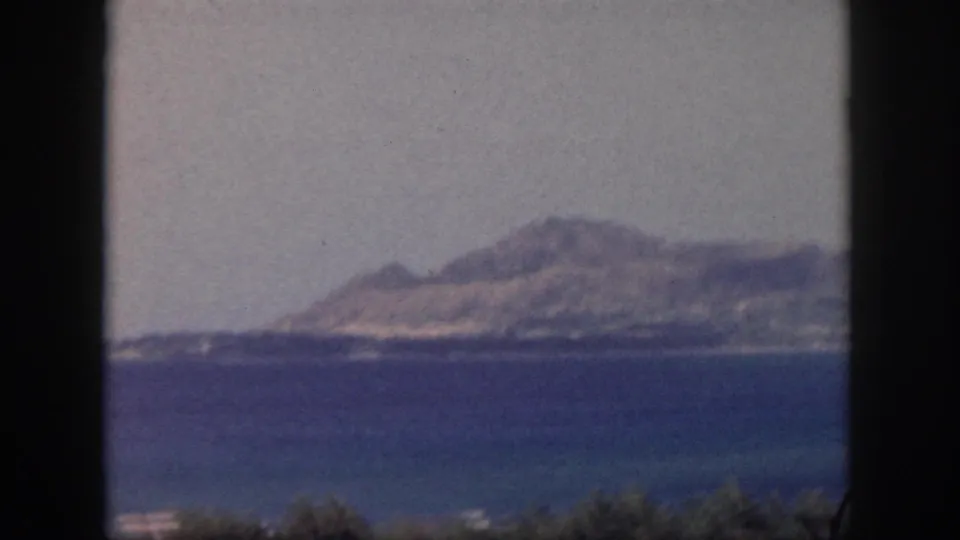 Oahu 1959 scenic frame showing blue bay water, a distant mountain ridge, hazy sky, and low vegetation at the bottom edge.