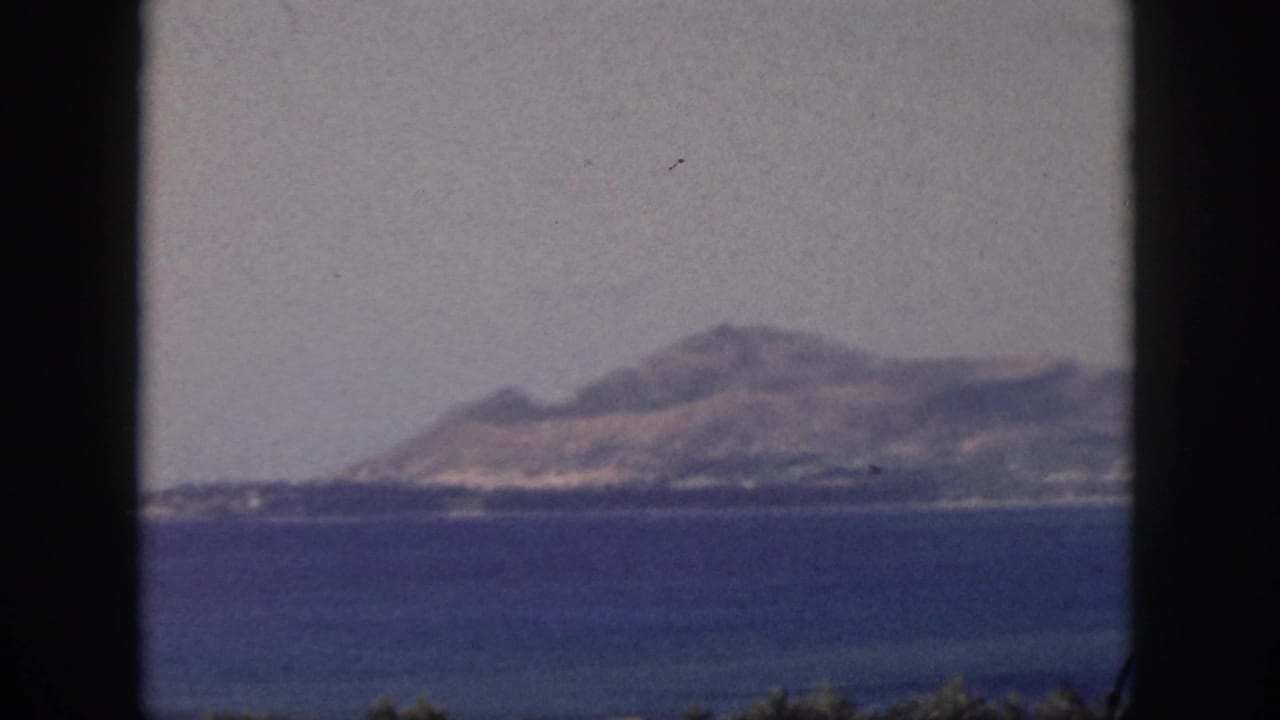 1959 Oahu home-movie frame showing a hazy island coastline beyond blue ocean with dark 8mm frame edges.