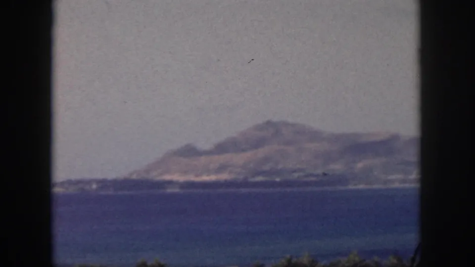 1959 Oahu home-movie frame showing a hazy island coastline beyond blue ocean with dark 8mm frame edges.