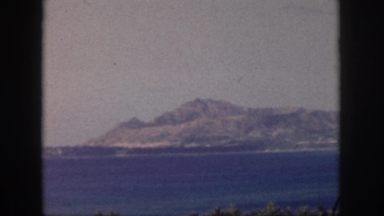 1959 Oahu seascape with a distant island shoreline, calm blue water, pale sky, and visible film-frame edges.