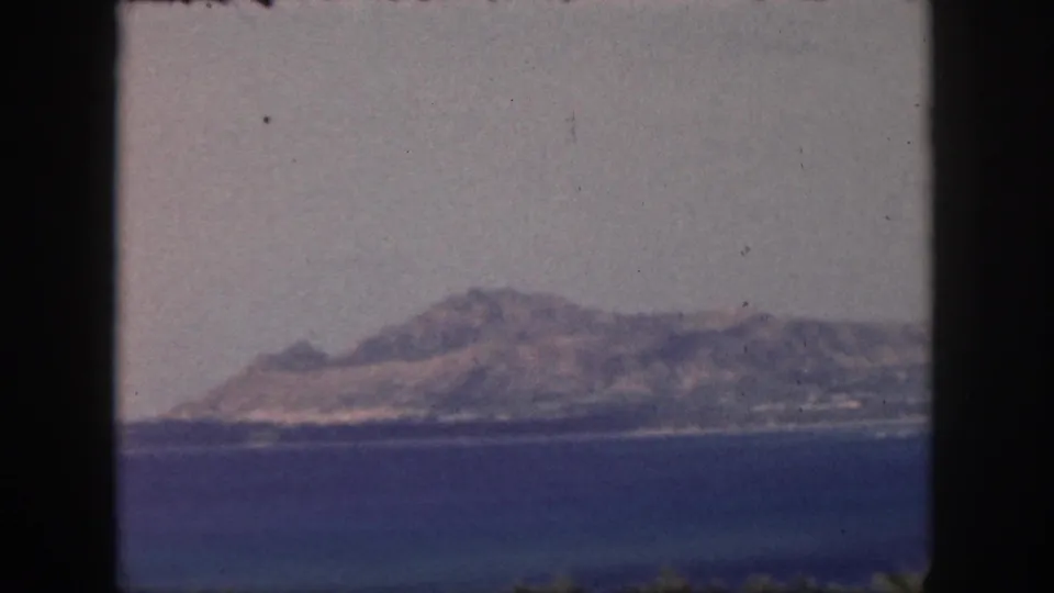 1959 Oahu 8mm frame showing a distant coast across blue water, with slight foreground foliage and film dust visible.