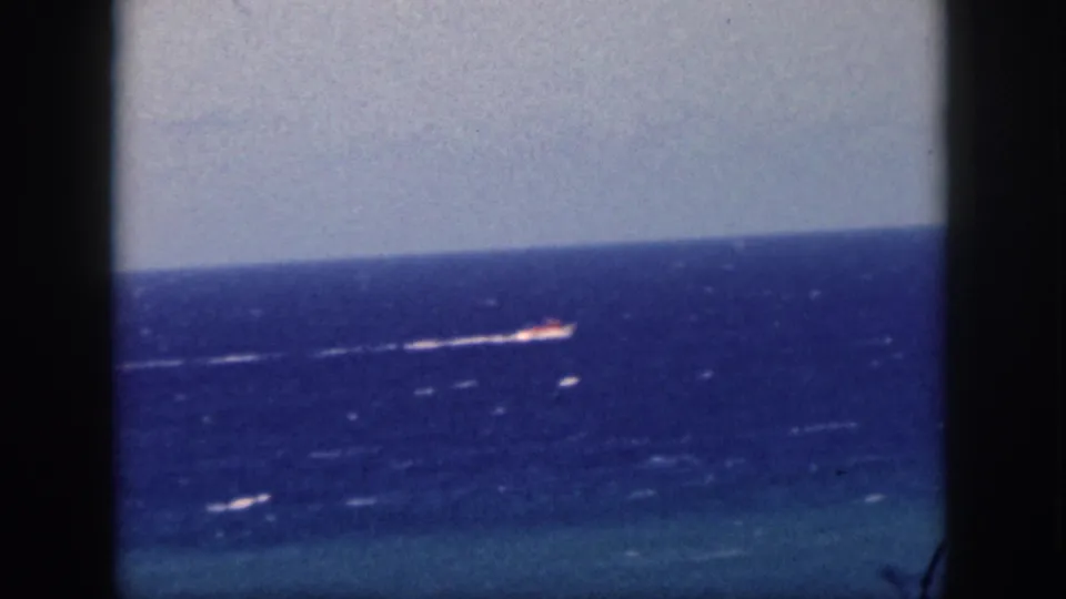 1959 Oahu home-movie frame showing a distant vessel crossing the Pacific, viewed through a dark cruise-window frame.