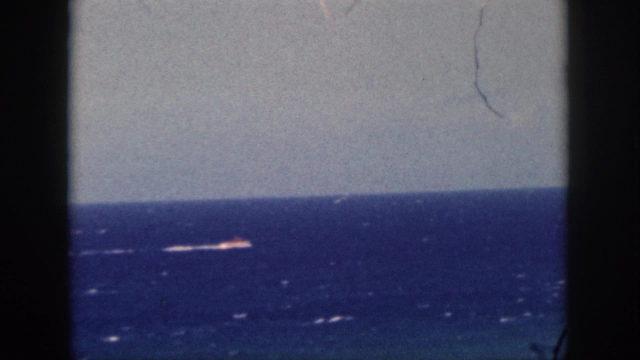1959 Oahu 8mm frame with a distant boat or bright wake on the ocean, framed by dark film edges and a pale sky.