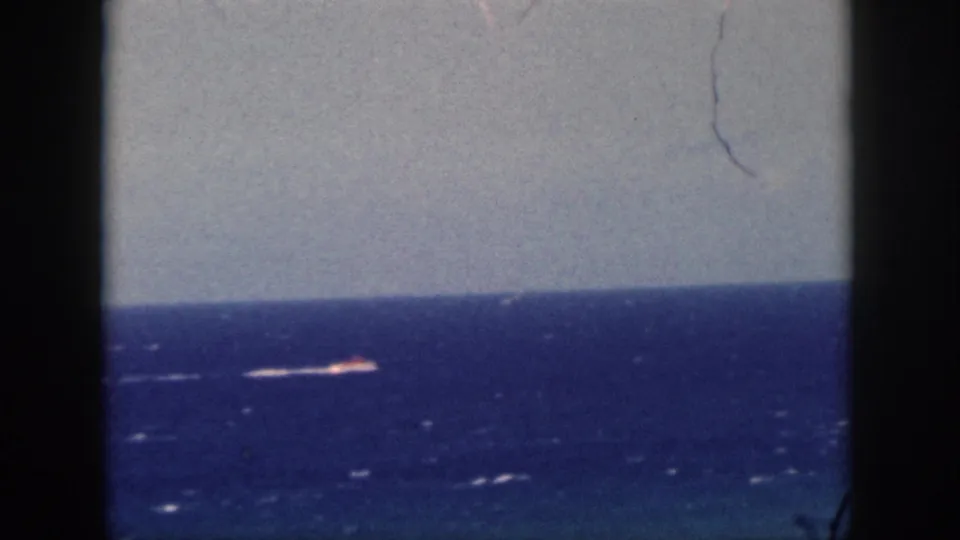 1959 Oahu 8mm frame with a distant boat or bright wake on the ocean, framed by dark film edges and a pale sky.