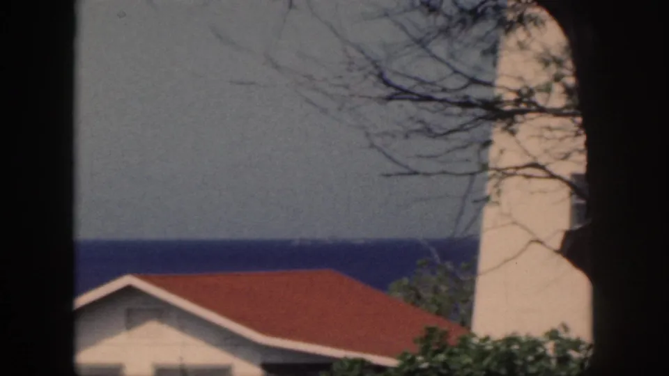 1959 Oahu home-movie frame showing a red-roofed coastal building and white lighthouse-like tower with ocean and branches behind it.