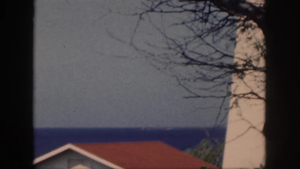 1959 Oahu shoreline frame with a white lighthouse-like structure, red roof, ocean horizon, and bare branches in faded 8mm color.