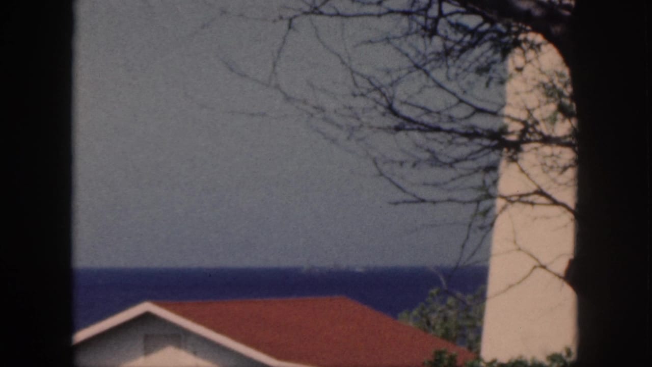 1959 Oahu 8mm frame showing a red-roofed coastal building, pale tower-like structure, tree branches, and blue ocean beyond.