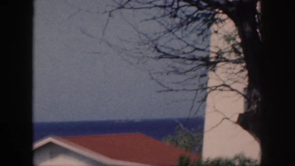 1959 Oahu 8mm frame showing a pale tower-like structure, red roof, foreground branches, and ocean under a hazy sky.