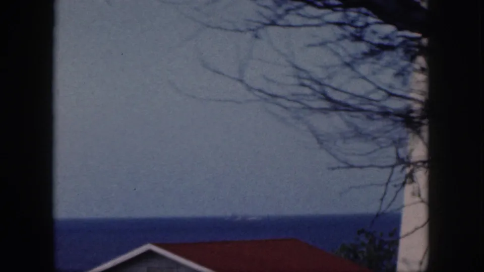 1959 Oahu 8mm frame showing a seaside view past dark framing, bare branches, a red roof, and the ocean horizon.