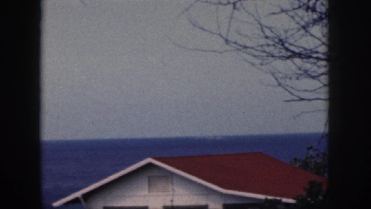 1959 Oahu 8mm frame showing a red-roof cottage and blue ocean seen through dark window mullions.