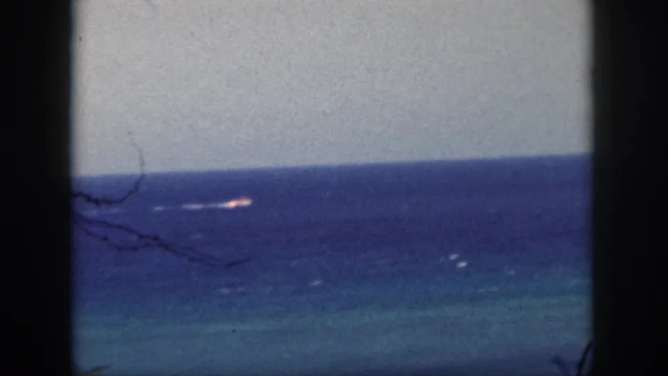 1959 Oahu coastal view with a small boat moving across deep blue water, partly framed by dark vertical edges and sparse branches.