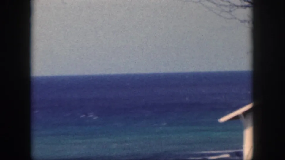 1959 Oahu shoreline frame with layered blue water, dark foreground framing, and the edge of a light-colored coastal building roof.