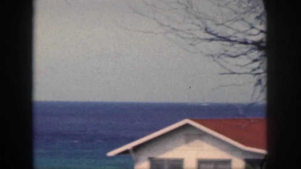 1959 Oahu 8mm frame showing a white coastal house with a red roof in front of layered blue ocean and sparse tree branches.
