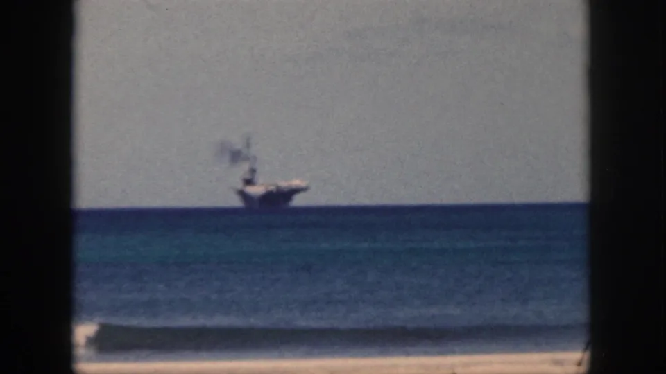 1959 Oahu home-movie frame with a distant aircraft carrier beyond blue ocean, low surf, and a pale sandy beach.
