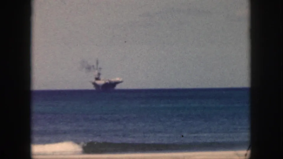 1959 Oahu shoreline frame showing a distant aircraft carrier on the horizon above calm surf and sand.