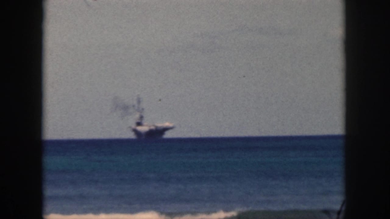 1959 Oahu 8mm frame showing a distant naval carrier at sea, viewed through dark vertical framing with smoke rising.