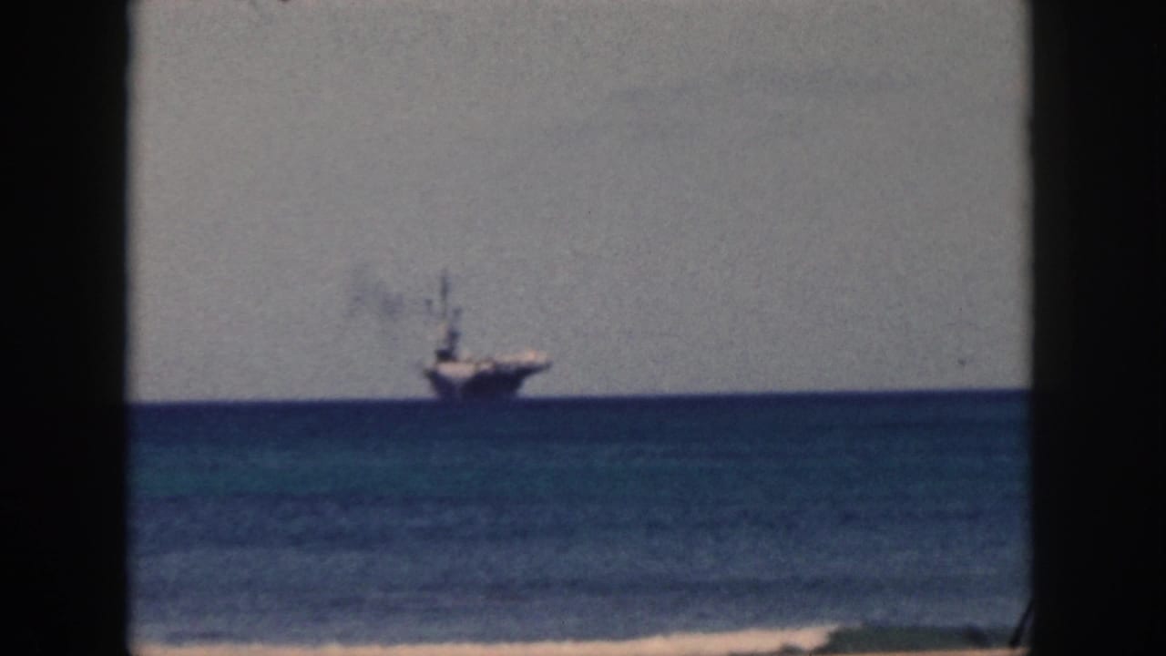1959 Oahu home-movie frame with a carrier on the horizon, smoke above the ship, and dark vertical borders in the foreground.