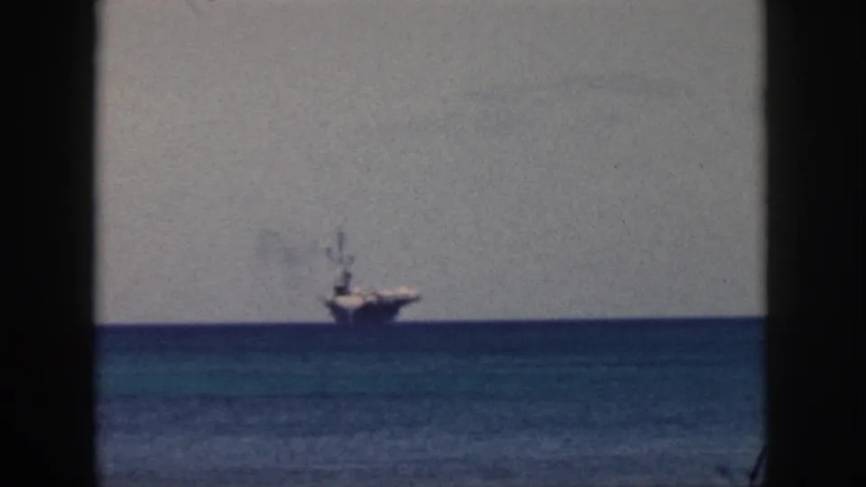 Distant 1959 Oahu view of a naval carrier offshore, with smoke visible above the vessel and soft 8mm grain across the sky.