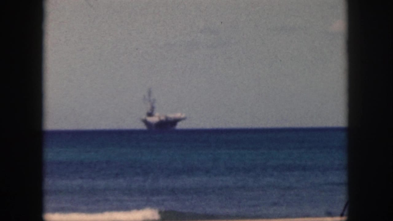1959 Oahu 8mm frame showing a distant aircraft carrier on calm water beyond a beach shoreline.