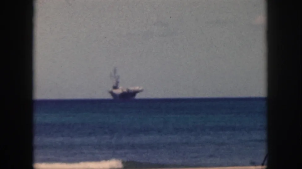 1959 Oahu 8mm frame showing a distant aircraft carrier on calm water beyond a beach shoreline.