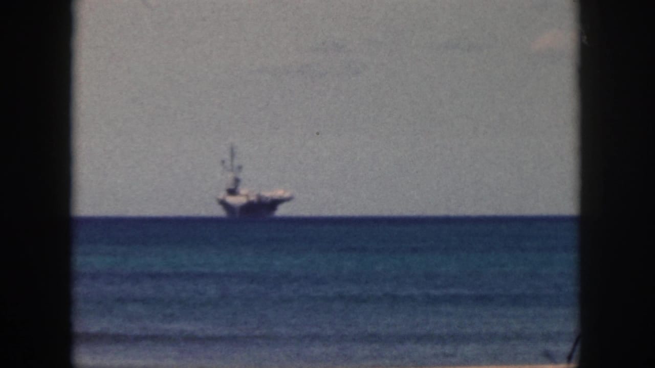 1959 Oahu 8mm frame showing a large distant naval-looking ship offshore across blue water, with a sandy beach edge and dark film gate borders.
