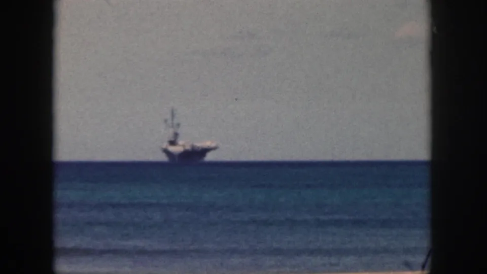 1959 Oahu 8mm frame showing a large distant naval-looking ship offshore across blue water, with a sandy beach edge and dark film gate borders.