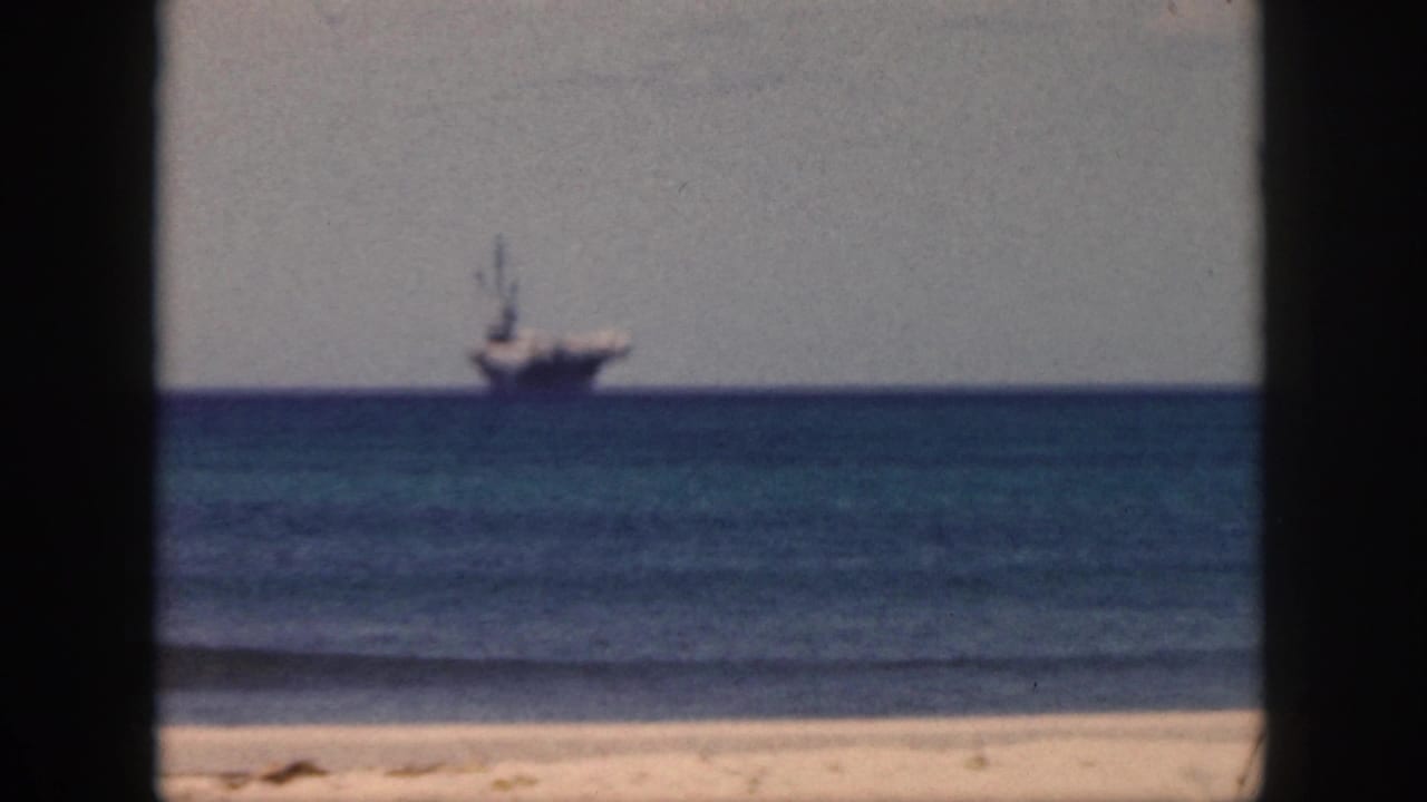 Distant large ship near the horizon over blue water, with beach sand at the bottom of the frame.