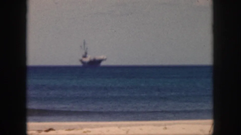 1959 Oahu home-movie frame with a large naval-looking vessel on the horizon, blue sea in the middle ground, and sand visible below.