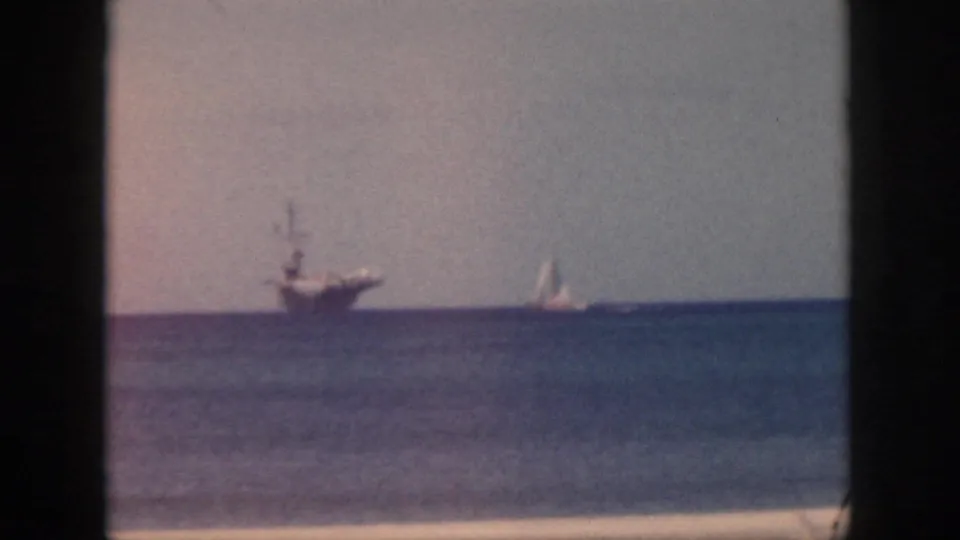 1959 Oahu frame showing a large offshore ship and a small white sailboat sharing the horizon beyond blue ocean and beach sand.