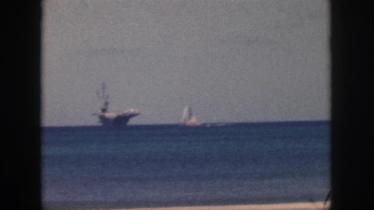 1959 Oahu home-movie frame showing a distant naval aircraft carrier, a small sailboat, calm blue water, gray sky, and dark window edges.