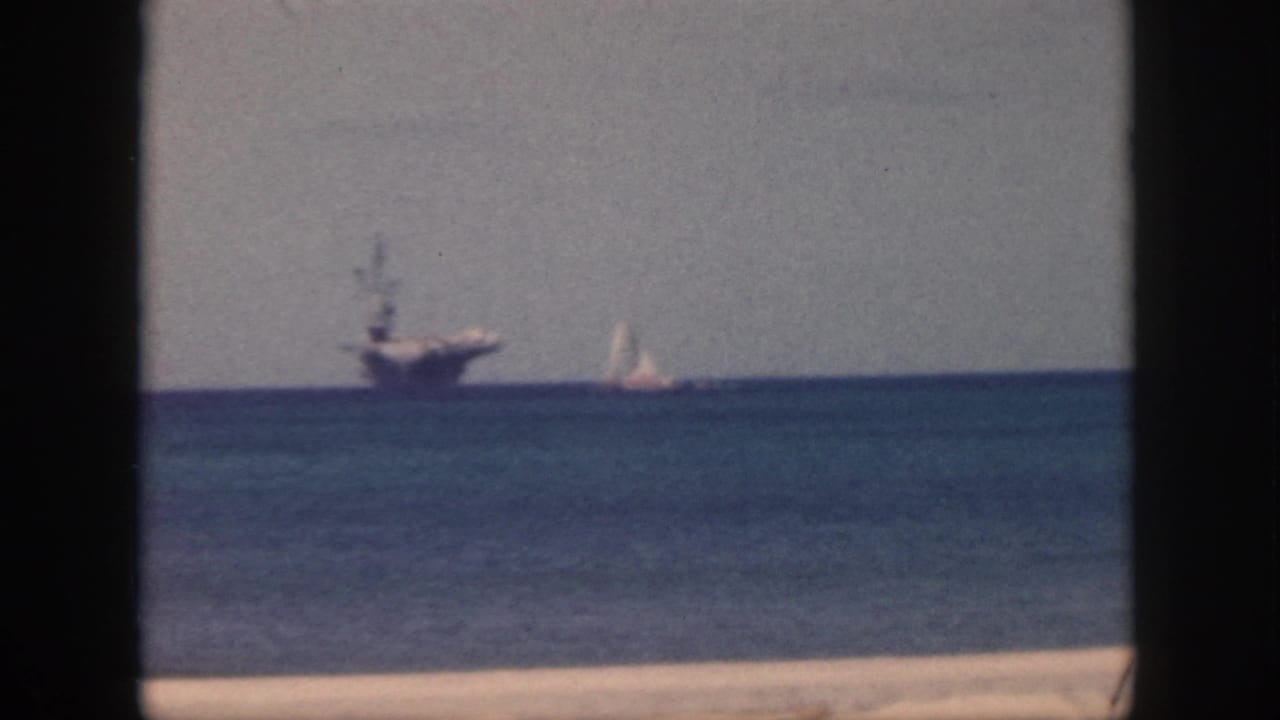Aircraft carrier and white sailboat near the horizon on calm water, framed by dark vertical bars.