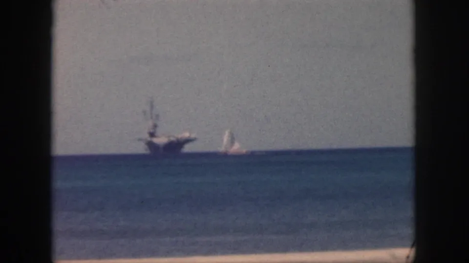 1959 Oahu 8mm frame showing a carrier and sailboat in the distance, calm ocean below, gray sky above, and a dark window edge at left.